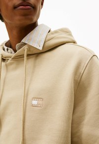 Beige Kapuzenpullover mit Kordeln, einer Fronttasche und einem kleinen beigen Logopatch. Über einem hellblau-weißen gestreiften Hemd mit Kragen getragen.