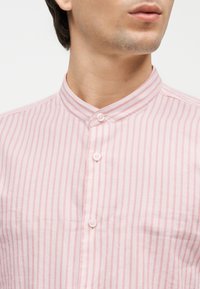 Άνδρας που φοράει πουκάμισο με κάθετες ριγέ ροζ και λευκές ρίγες, με γιακά Μανδαρίνου, κουμπωμένο στο λαιμό και κατά μήκος του εμπρός μέρους.