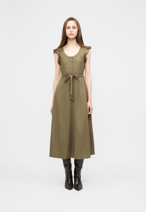 Vrouw draagt een olijfgroene midi-jurk met korte mouwen, een geknoopte top, een gestrikte taille en zwarte knielaarzen, staand tegen een effen achtergrond.