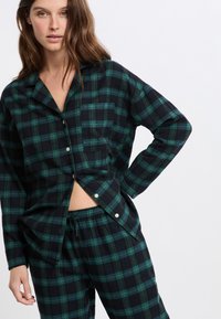 Kobieta w ciemnozielonej i czarnej koszuli nocnej w kratę z guzikami oraz pasujących spodniach z elastycznym pasem, stojąca na tle białej ściany.
