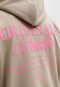 Sweat à capuche beige avec un grand texte rose indiquant « ORIGINALS Studios » et de plus petites phrases imprimées en rose au dos.