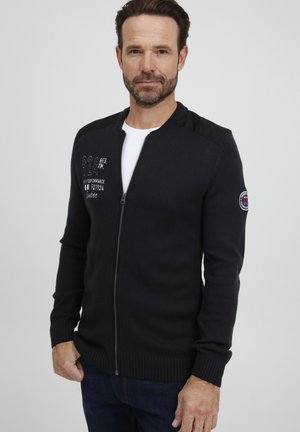 Suéter navy de cremallera con dobladillo y puños acanalados, con acentos de cuero negro en los hombros y detalle bordado en blanco en el pecho.