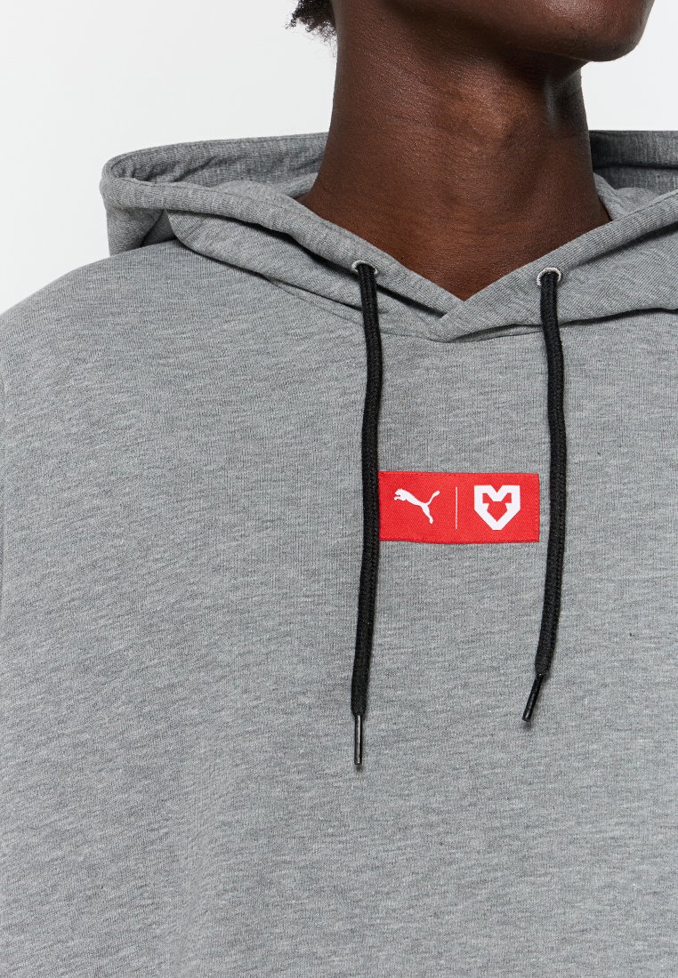 Acercamiento a una persona que lleva puesta una sudadera gris con cordones negros y un parche rojo con el logotipo de Puma y un símbolo en forma de M.