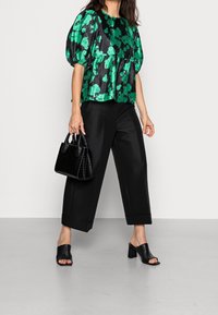 Donna in pantaloni ampi neri a vita alta, scarpe con tacco a blocco aperte nere, top a maniche puff floreale verde e nero, che tiene in mano una borsa in pelle nera.