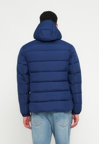 Mørkeblå dunjakke med hætte, der har horisontale quiltede sektioner og elastiske manchetter, parret med lyseblå denimjeans.