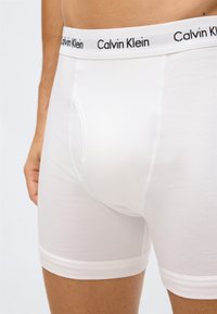 Detailní záběr na osobu v bílých boxerkách Calvin Klein s elastickým páskem s logem.