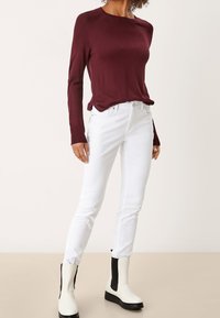 Maglione bordeaux a maniche lunghe, jeans bianchi slim-fit e stivaletti bianchi con accenti neri, dotati di polsini a costine e design aderente.