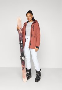 Εξοπλισμός σκι: μια γυναίκα κρατάει ένα ροζ σκί με μαύρους δεσίματα, φορώντας κόκκινο αδιάβροχο μπουφάν, γκρι μπλούζα, λευκό παντελόνι και μαύρα παπούτσια σκι.