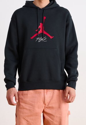 Osoba nosząca czarną bluzę z kapturem z czerwonym logo Jumpman i napisem „Flight”, w połączeniu z jasnopomarańczowymi spodniami.