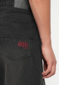 Črne jeans hlače z zadnjim žepom, na katerem je rdeče vezeno besedilo "REDEFINED REBEL RODEO." Usnjen žepni znak z logotipom nad žepom.