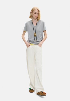 Młoda osoba stoi z rękami w kieszeniach, ubrana w szarą koszulkę z dekoltem w serek, szerokie białe spodnie, brązowe mokasyny i długi naszyjnik z zawieszką.