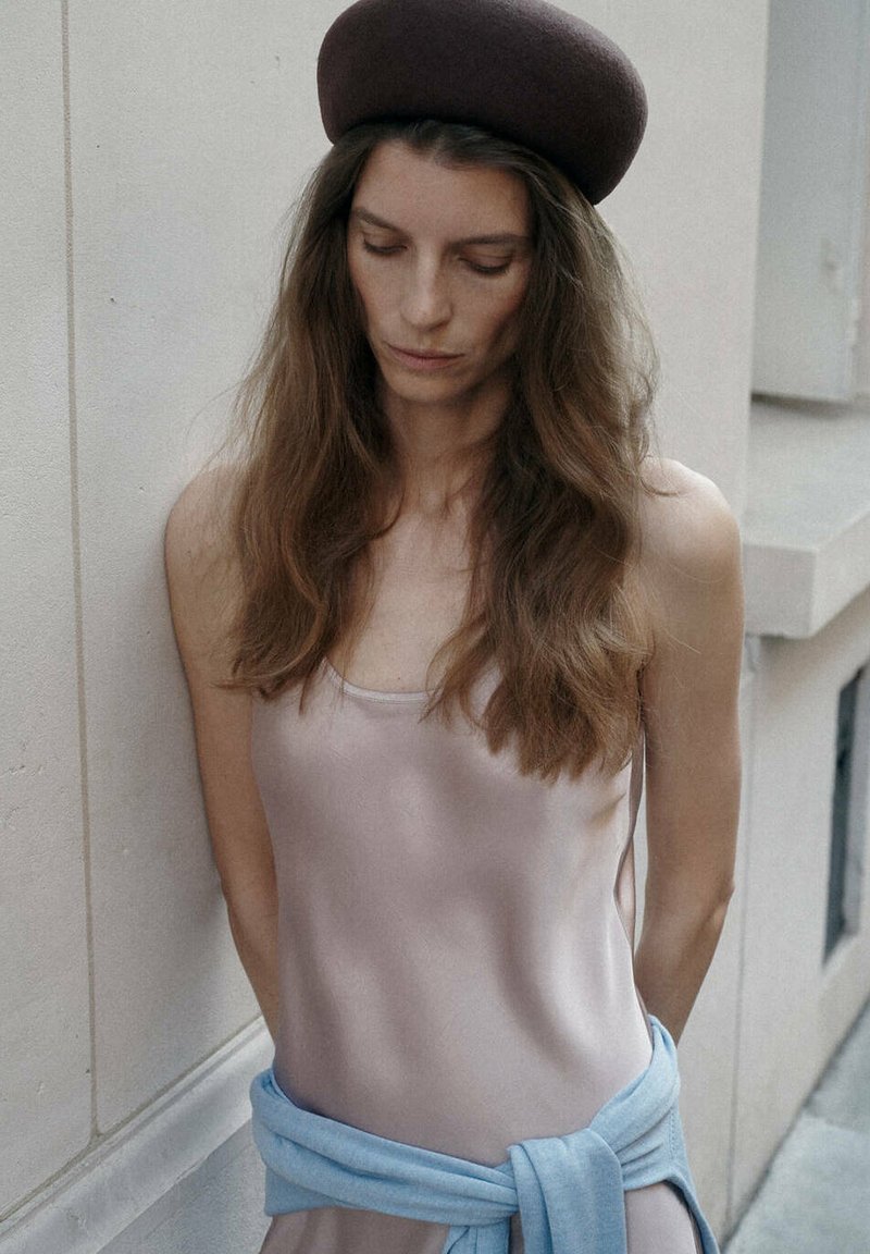 Jeune femme aux longs cheveux portant un béret marron, un haut sans manches rose clair et un pull bleu clair noué autour de la taille, debout près d’un mur.