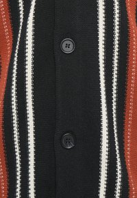 Gebreide cardigan met zwarte, witte en roestkleurige strepen. Zwarte knopen zijn zichtbaar, wat de textuur en het kleurcontrast in de stof benadrukt.