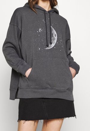 Sweat-shirt gris avec une poche frontale, présentant un graphisme de lune et des motifs tachetés. La coupe est ample avec un ourlet et des poignets côtelés. Porté avec une jupe noire.