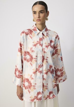 Žena koja nosi široku košulju dugih rukava s gumbima, s uzorkom crvenih, bež i plavih tie-dye cvjetnih motiva te zlatne viseće naušnice, na bijeloj pozadini.