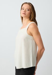 Top senza maniche beige chiaro con scollo a quadrato, caratterizzato da una texture liscia e un design semplice, abbinato a pantaloni scuri.
