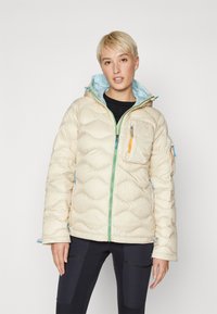 Beige pufferjack met een blauwe binnenvoering. Heeft golvenpatronen, groene en oranje ritsen, en meerdere zakken. Zacht, gestructureerd materiaal.