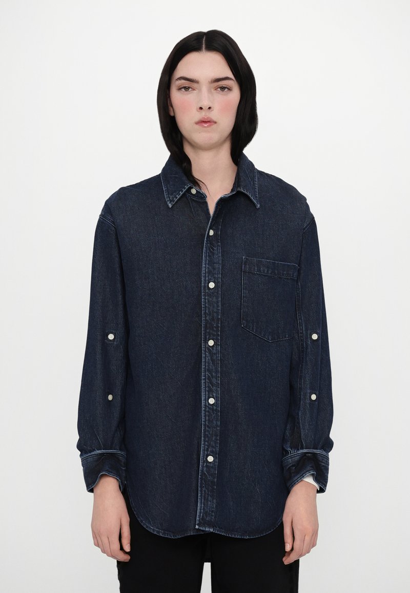 Chemise en denim bleu foncé avec un col pointu, poche avant, poignets à boutons et ourlet arrondi. Texture douce avec des détails de couture visibles.