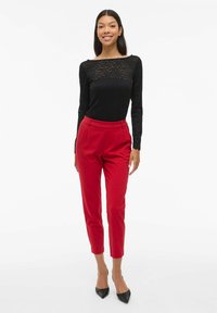 Haut noir à manches longues avec un design en tricot ouvert, associé à un pantalon rouge ajusté. Le mannequin porte des escarpins noirs à bout pointu, se tenant sur un fond blanc.