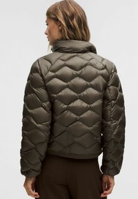 Kobieta z włosami do ramion, ubrana w ciemnooliwkową pikowaną kurtkę puchową, widziana z tyłu na jednolitym tle.
