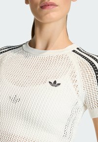Top deportivo blanco de punto con diseño de malla transpirable. Presenta las rayas negras de Adidas en las mangas y el logo en el pecho. Cuello redondo sin costuras.