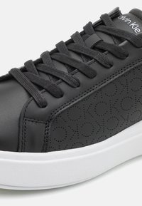 Black sneaker with smooth leather upper featuring embossed logo pattern, black laces, and a white rubber sole.