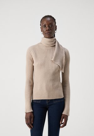 Maglione beige a collo alto con dettaglio drappeggiato. Maniche lunghe, tessuto strutturato e silhouette aderente. Abbinato a jeans scuri.