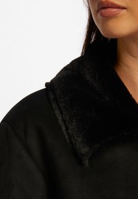 Svart frakk med en teksturert, myk krage. Stoffet ser ut til å være mykt med en glatt, matt finish. Nærbilde av krage og halslinjedetaljer.