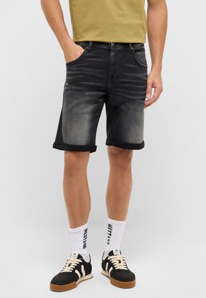 Man wearing faded black rolled-hem denim shorts, beige t-shirt, white crew socks with text, and black and beige sneakers, standing against a plain background.