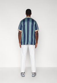 Rövid ujjú ing, függőleges kék és zöld csíkokkal, galléros nyakkivágással krémszínű díszítéssel, fehér nadrággal és szürke tornacipővel viselve.