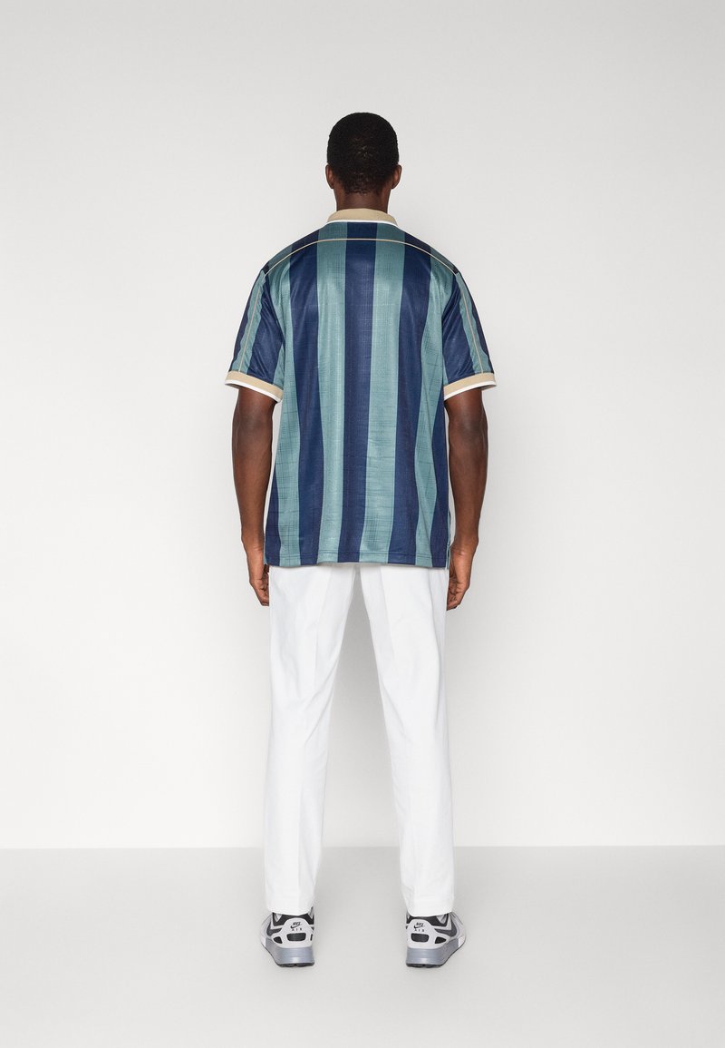 Camicia a maniche corte con righe verticali blu e verdi, collo con dettagli color crema, abbinata a pantaloni bianchi e sneaker grigie.