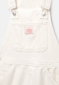 Salopette in denim bianco con clip metalliche regolabili, tasca sul petto con etichetta stampata rosa e tasche laterali. Tessuto testurizzato con dettagli di cucitura.