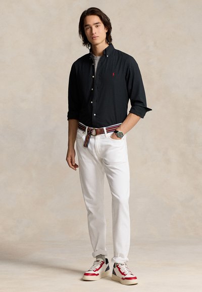 Mlad moški, oblečen v črno srajco na gumbe, bele hlače s črtastim pasom in rdeče, bele ter mornarsko modre superge, stoji z eno roko v žepu.