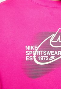 Pink Nike t-shirt featuring white branding text, circular design with striped pattern, and crew neckline. Soft cotton fabric.