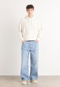 Felpa crema con tasca frontale e cordini abbinata a jeans a gamba larga blu chiaro e scarpe beige, su sfondo semplice.