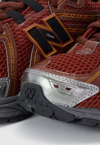 Scarpe atletiche in rete rossa con accenti marroni e dettagli metallici argentati. Presentano lacci neri e suola in gomma con logo. Design ventilato.