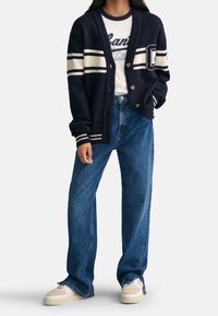 Granatowy kardigan w kremowe paski, z guzikami i ściągaczami przy rękawach, noszony na białej koszuli z nadrukiem. W zestawieniu z luźnymi niebieskimi dżinsami i sneakerami.