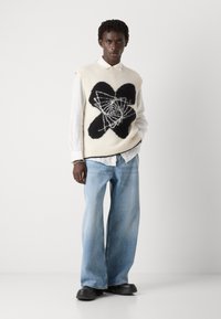 Chaleco de punto en color crema con un gran diseño gráfico en negro, superpuesto a una camisa blanca con cuello, combinado con jeans azules holgados y zapatos negros.