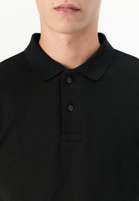 Persoană îmbrăcată cu un tricou polo texturat negru, având două butoane negre închise la guler, lăsând să se vadă gâtul și partea inferioară a feței.