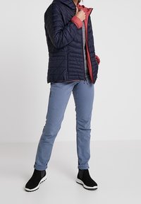 Marineblauer gesteppter Mantel mit rotem Innenfutter, darunter ein graues Hemd, blaue Hose und schwarze Stiefeletten mit weißen Sohlen. Kapuzen-Design.