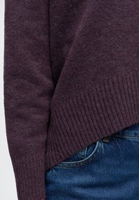 Gros plan d’une personne portant un pull tricoté violet foncé et un jean bleu en denim, montrant la manche du pull et l’ourlet côtelé.