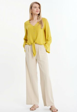 Blusa gialla a maniche lunghe con dettaglio a fiocco frontale, piccoli bottoni, abbinata a pantaloni beige a gamba larga e sandali beige. Tessuto di trama liscia.