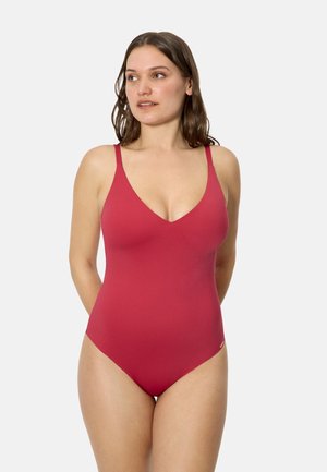 Mujer con cabello castaño que lleva un traje de baño entero rojo sin mangas, de pie contra un fondo claro y liso, mirando hacia un lado.