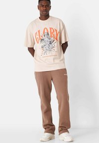 Beige grafisk t-shirt med "GLORY" i orange, med en illustration. Kompletteras av bruna sweatpants och vita sneakers.