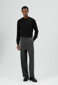 Zwarte lange mouwen shirt gecombineerd met grijze, op maat gemaakte broek met rechte pijpen. Model draagt zwarte schoenen; minimalistische achtergrond.