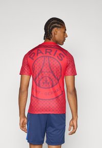 Rød PSG-trøje med et geometrisk mønster, med "PARIS" og et logo i mørkeblå. Båret med marineblå shorts, der fremhæver det atletiske design.