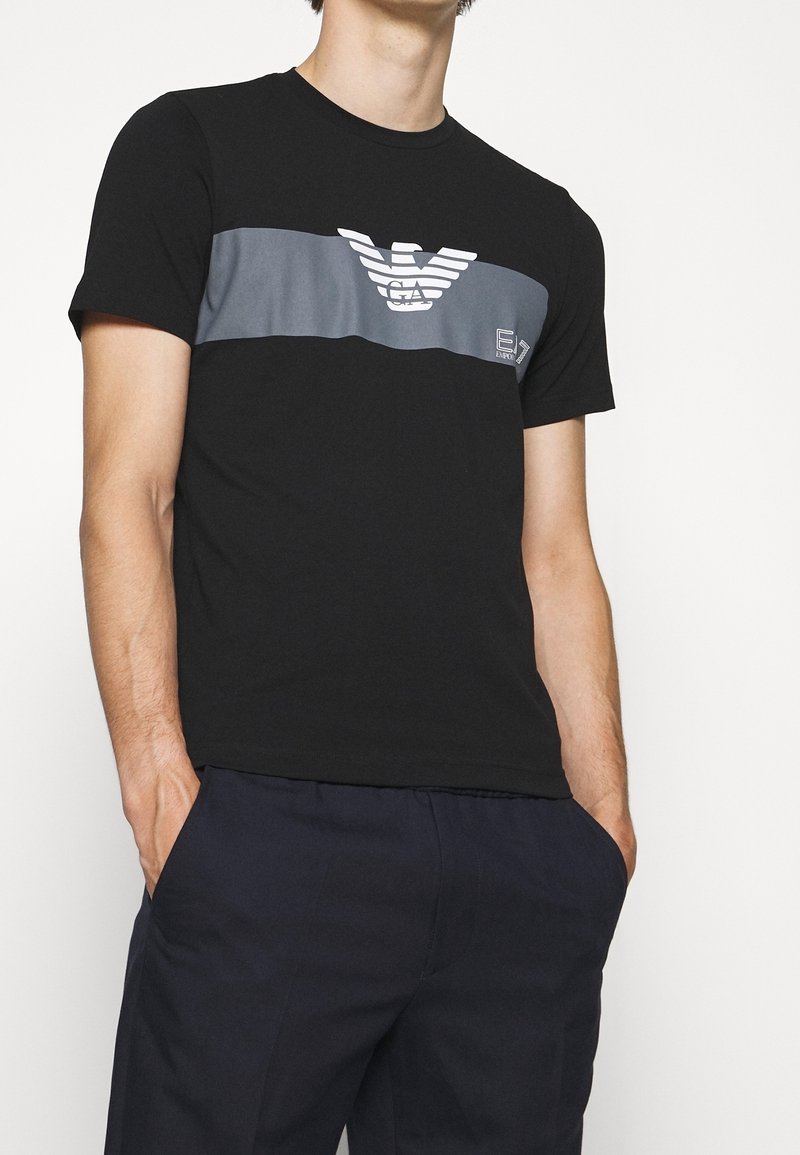 Schwarzes T-Shirt mit einem grauen horizontalen Streifen, der ein Logo trägt. Kurze Ärmel, Rundhalsausschnitt, aus glattem Stoff gefertigt. Legere Passform.