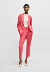 Helderroze op maat gemaakte blazer en bijpassende broek met een riem. Witte, getailleerde top eronder. Puntige, crèmekleurige muiltjes maken de outfit compleet.