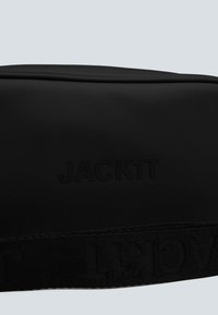 Černá taštička vyrobená z texturovaného materiálu s vyraženým logem "JACK1T" na přední straně a vzorovaným tkaninovým popruhem pod spodním okrajem.
