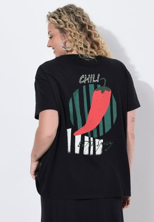 Frau mit lockigem blondem Haar, die ein schwarzes T-Shirt mit rotem Chili und dem Text "Chili extra spicy" auf dem Rücken trägt.